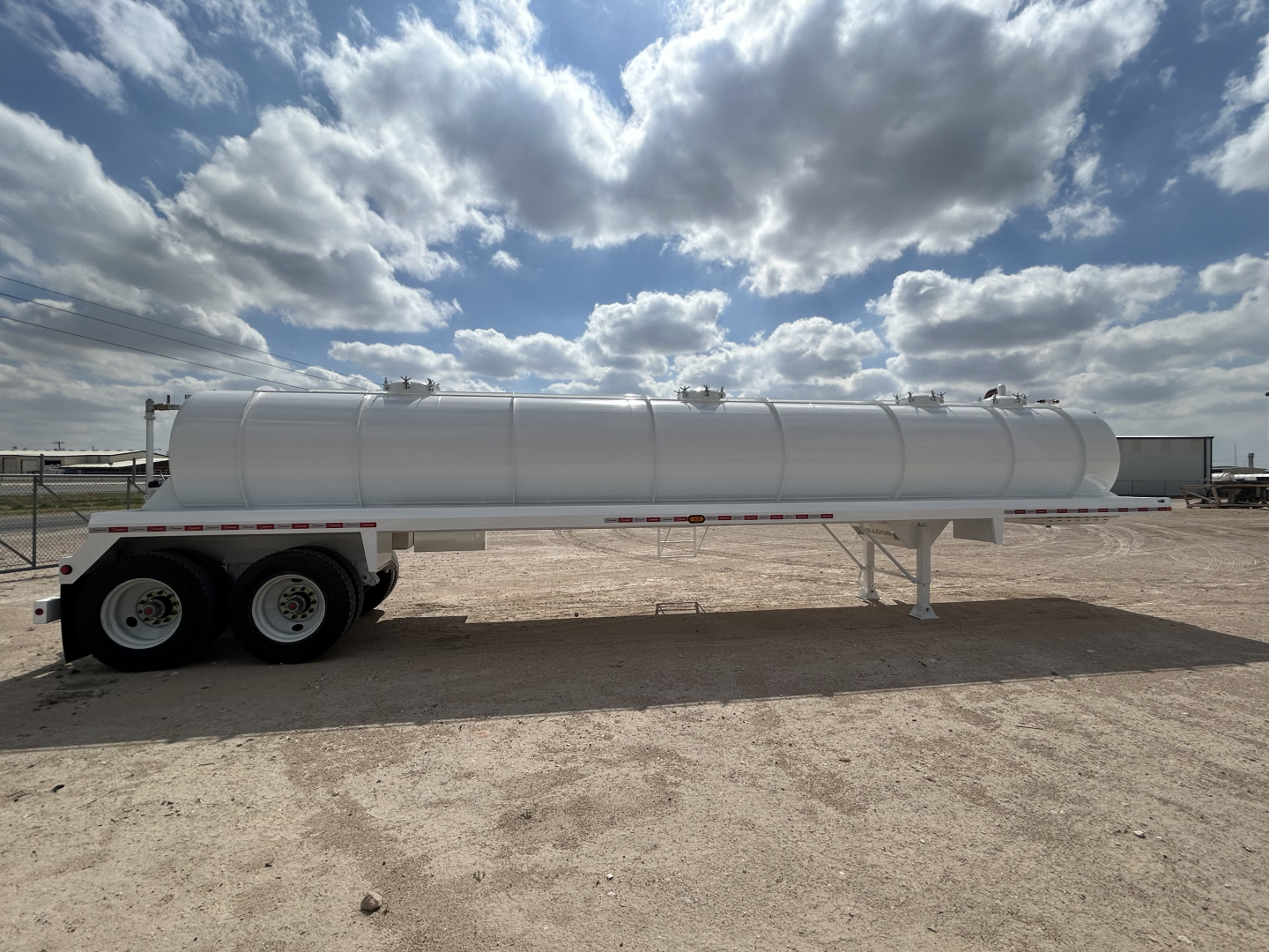 Trailer-mounted vacuum tanker cylindrical tank on dual-axle trailer, white finish, side view with support legs, bright sky with clouds.