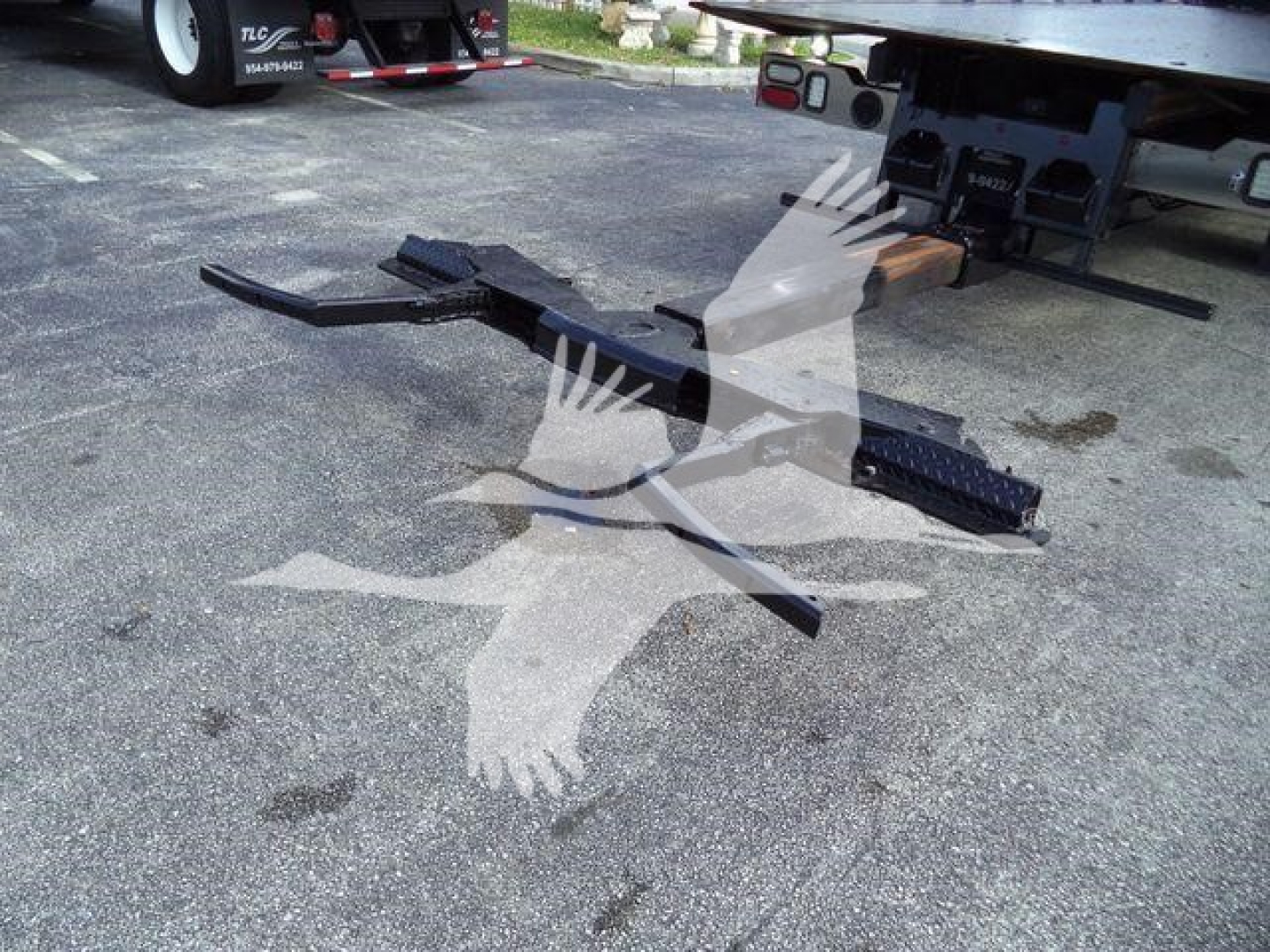 Truck rollback equipment showing a black metal hitch assembly and crossbar lying on pavement near a Ford F-Series 2022 F600 in a lot.