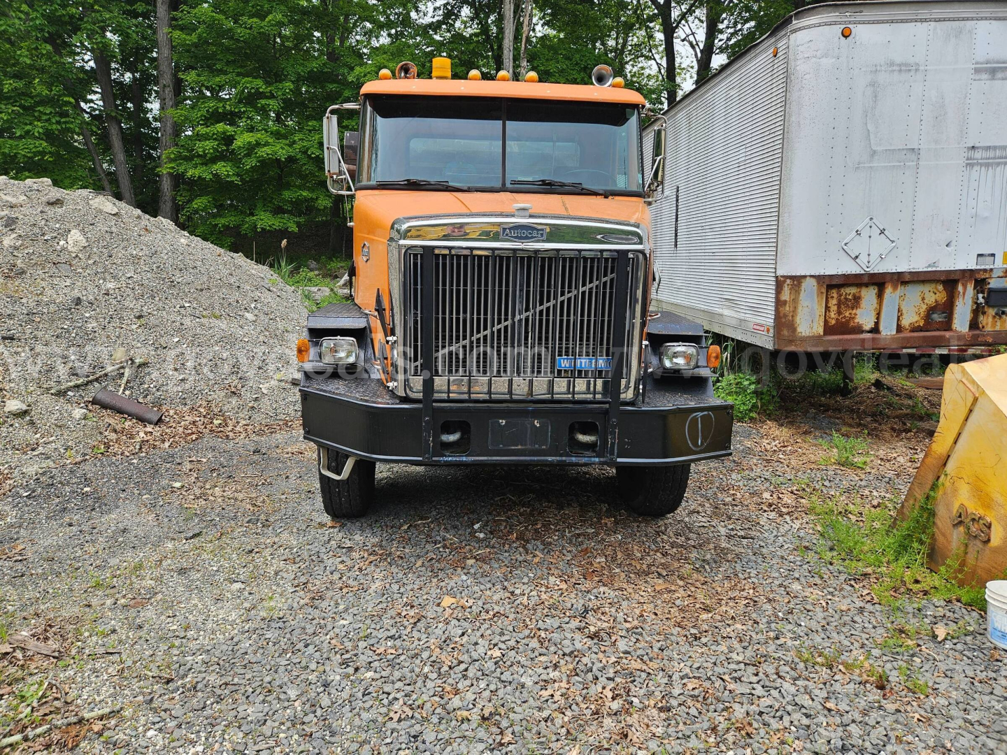 Orange Autocar truck from front view in a yard, heavy-duty grille, chrome bumper, large black tires, with a white trailer on the right and pile of gravel on left.