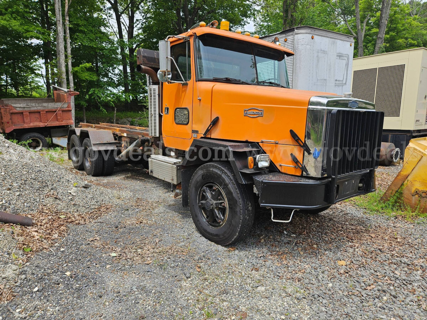 Autocar ACL roll-off truck in orange with black front bumper and grille, side exhaust, large cab, on a gravel lot with equipment in background.