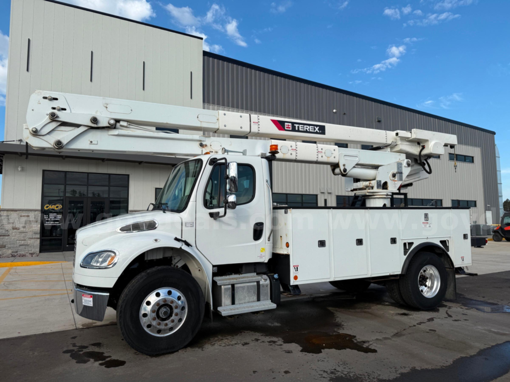 Truck bucket lift: Freightliner Terex TC-55 utility body with boom elevated, white cab, service compartments along side, dealership building in background.