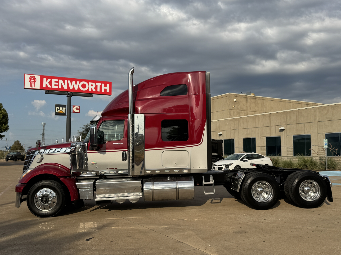 Truck side view showing a red and white 2021 International Lonestar sleeper cab with dual rear axles and Kenworth dealership signage in background.