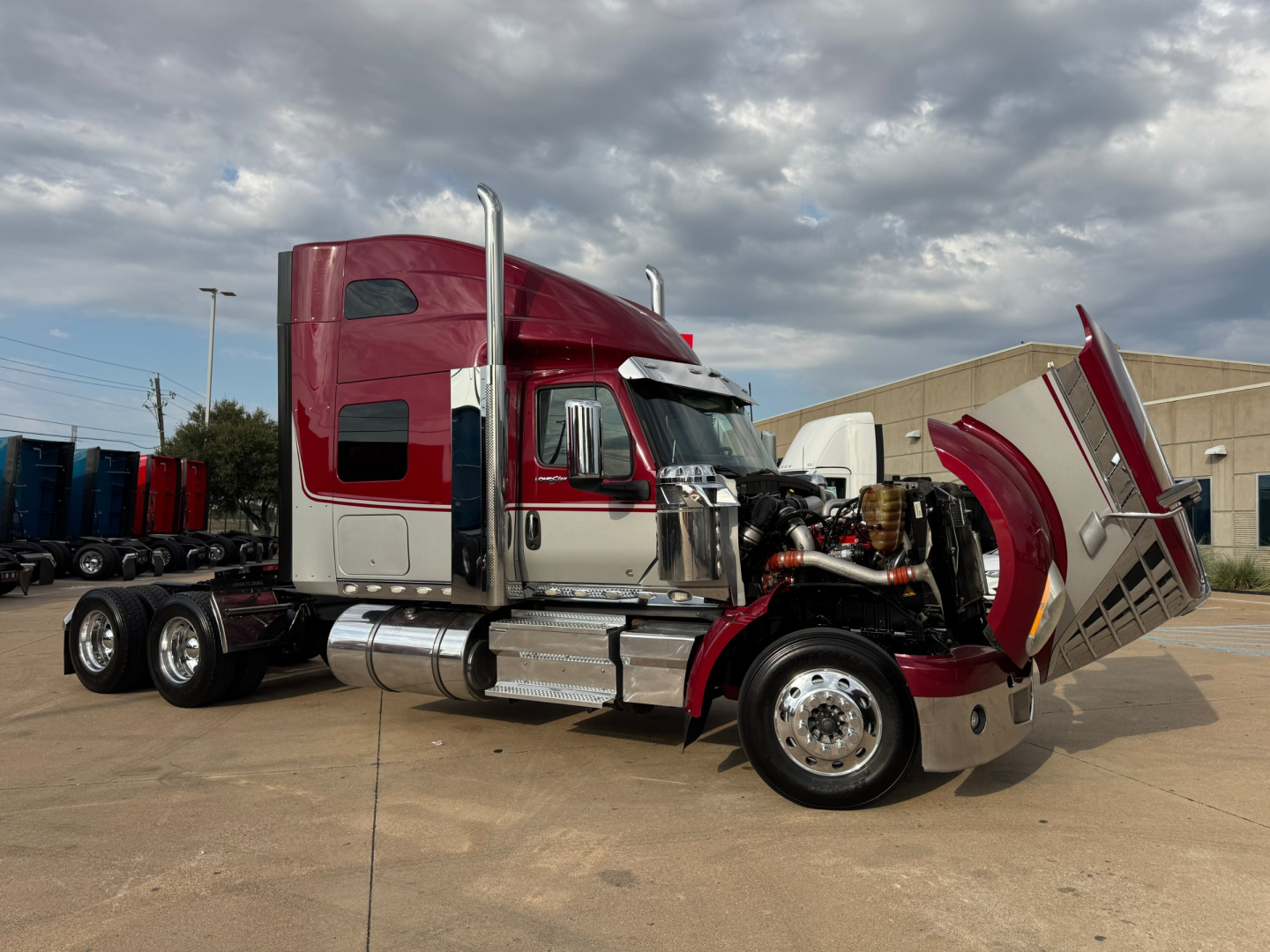 International Lonestar truck with raised hood showing engine bay and chrome exhaust stacks, maroon and silver cab, tandem rear axles, on concrete lot.
