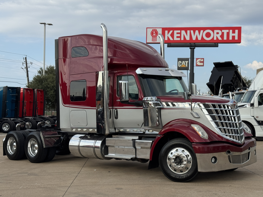 Truck cab in conjoined sleeper tractor configuration with maroon and silver color showing chrome exhaust stacks and chrome bumper, Kenworth dealership sign in background.