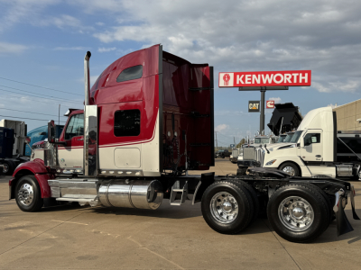 Truck cab and chassis with a Kenworth sign in background, red sleeper cab, chrome fuel tanks, dual rear axles.