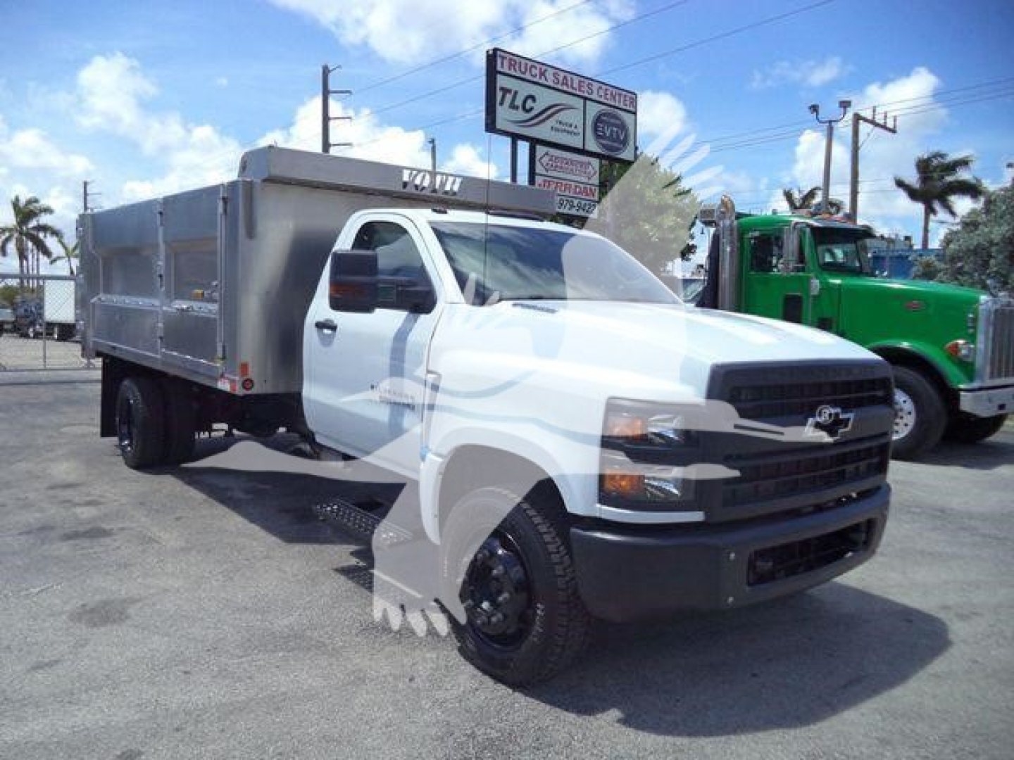 Dump truck with aluminum cargo bed and white cab, Chevrolet grille, in a dealership lot. Visible text on signage in background and truck body.
