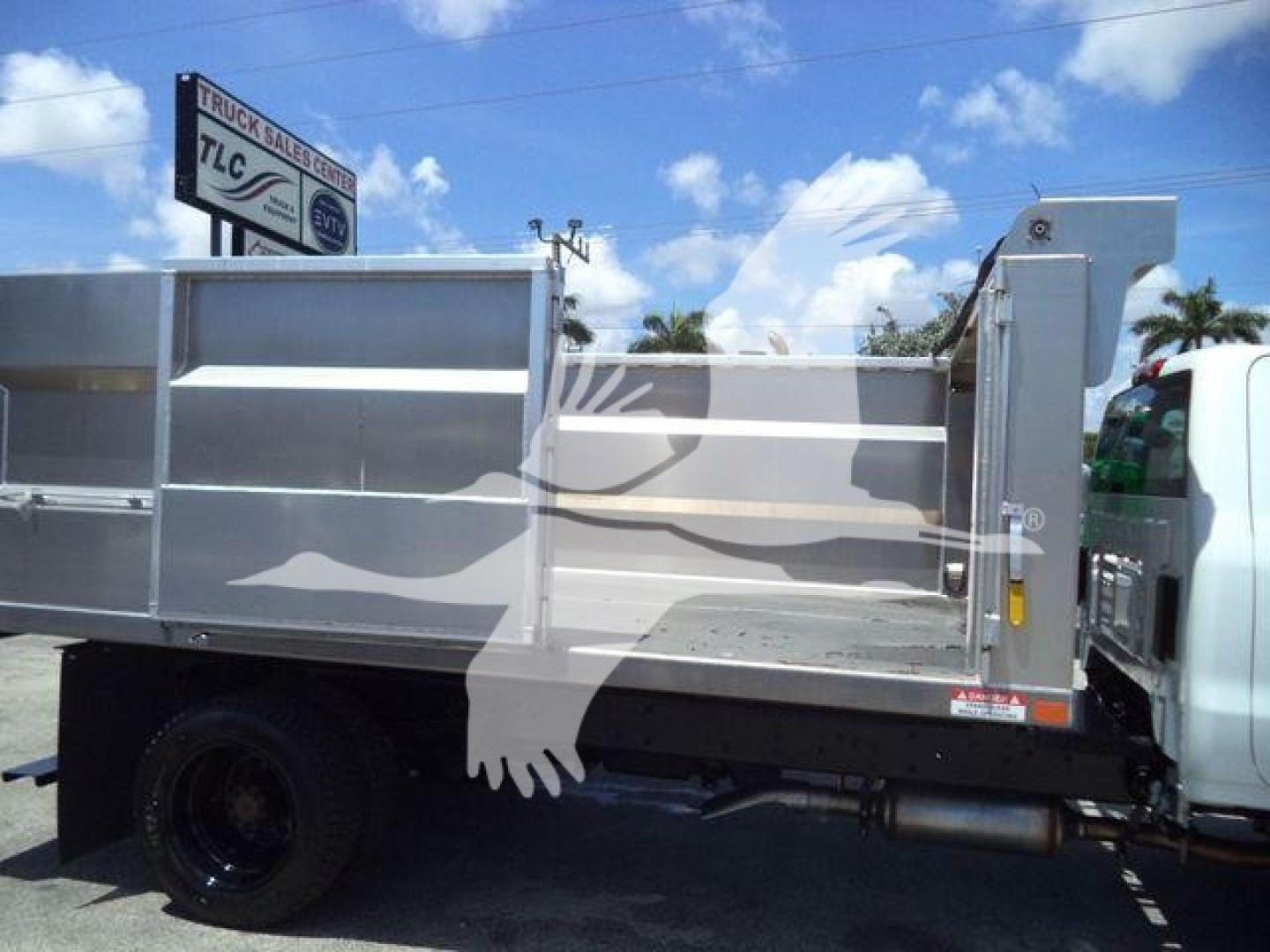 Dump truck bed side view showing aluminum dump body on a Chevrolet Silverado 6500HD chassis with branding and a stock sign in the background.