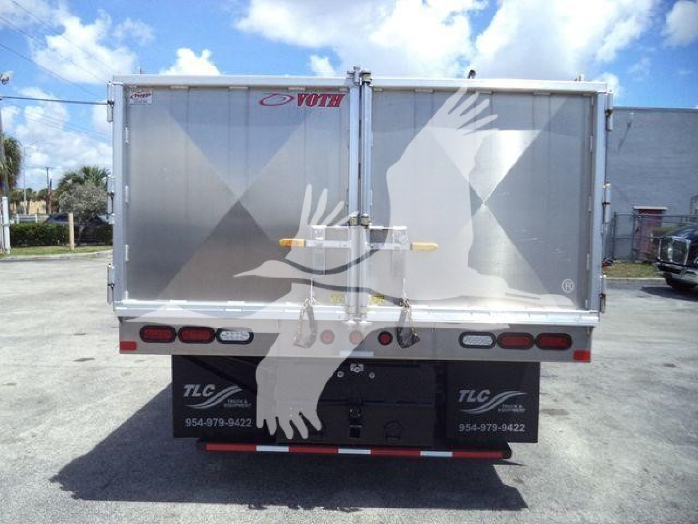 Rear view of a Chevrolet Silverado 6500HD dump truck with silver dump bed, taillights, and bumper. Brand logos TLC and VOTH on the bed doors.