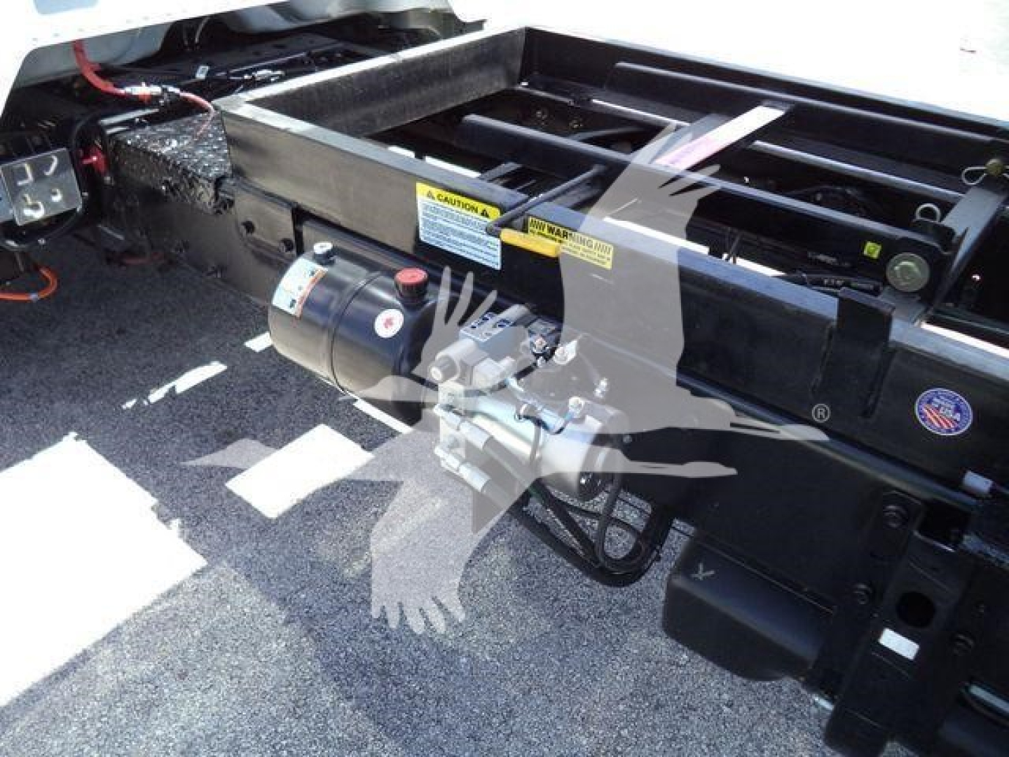 Truck chassis with label warnings and a hydraulic pump assembly mounted on the frame; visible yellow warning stickers and a circular UK flag-style sticker.