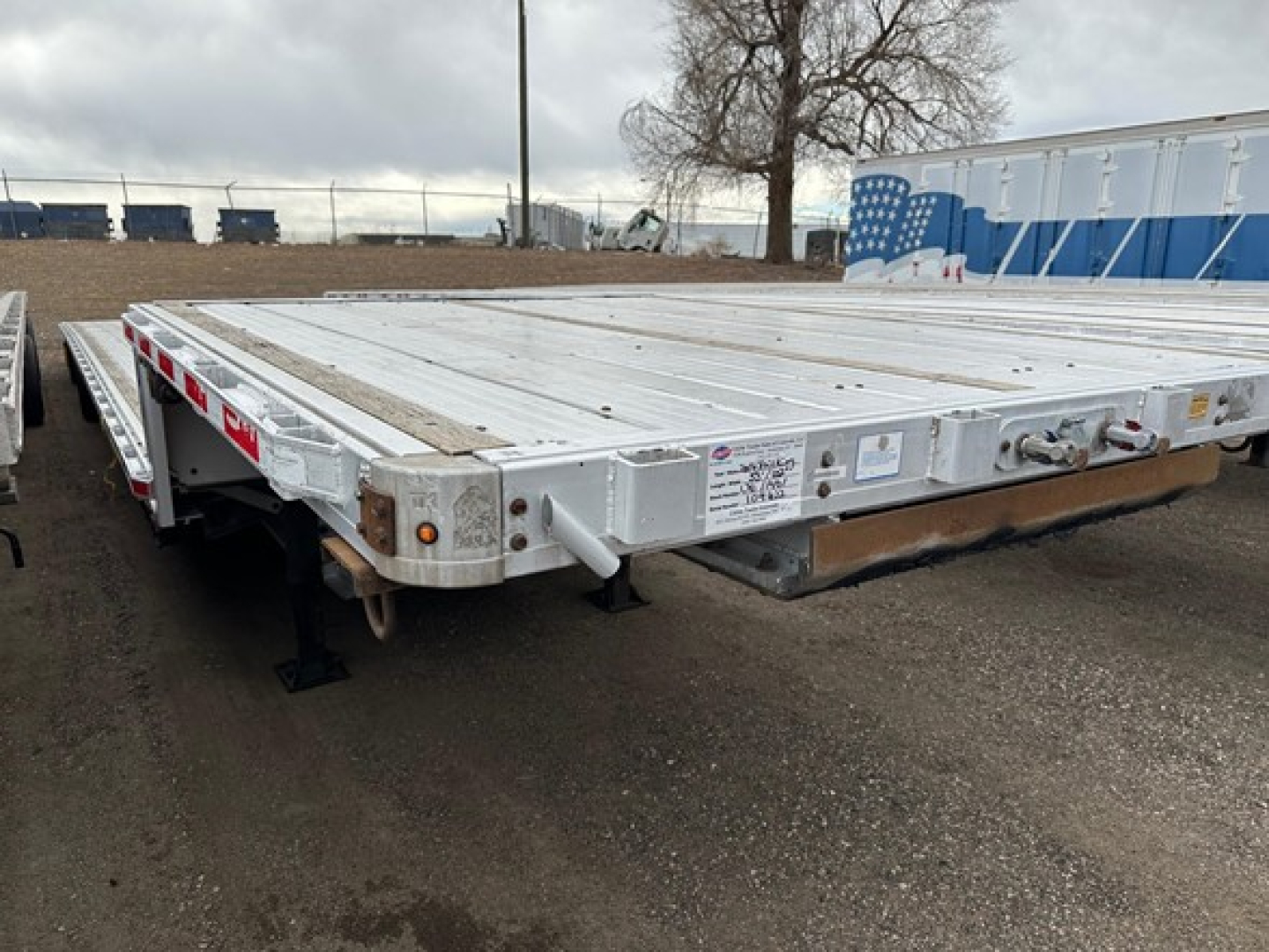 Flatbed trailer deck showing aluminum construction with drop-down ramps and side safety rails. Visible labels/stickers on front corner with serial-like text. No clear readable numbers due to distance.