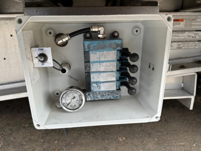 Valve/control block with multiple hydraulic lines and gauge inside a white junction box on a drop-deck trailer.