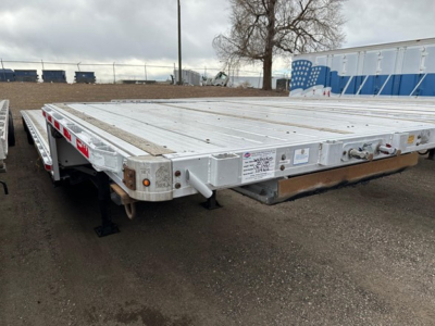 Flatbed trailer deck showing aluminum construction with drop-down ramps and side safety rails. Visible labels/stickers on front corner with serial-like text. No clear readable numbers due to distance.
