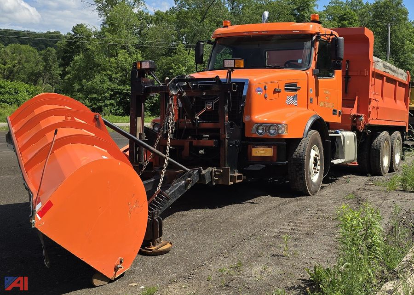 Orange Volvo VHD dump truck with large orange snow plow attachment in front, side view parked on dirt with trees in background.
