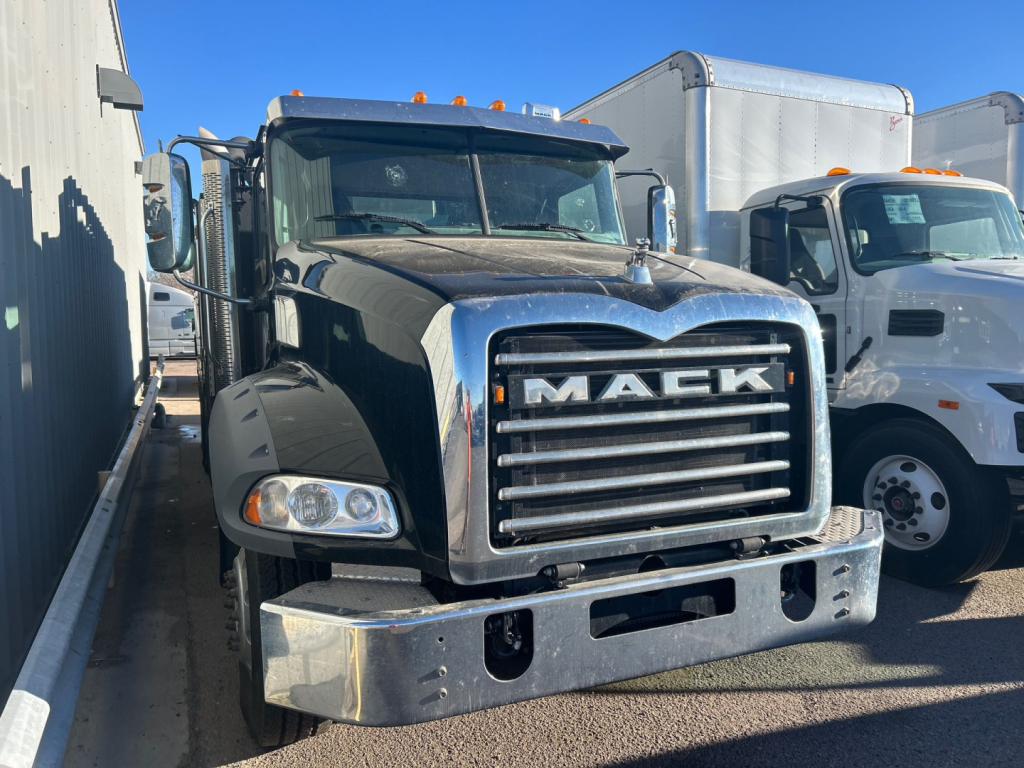 Front view of a black 2023 Mack GR64B truck in a lot showing the large chrome grille with MACK emblem, bumper, and headlights; other commercial trucks in background.