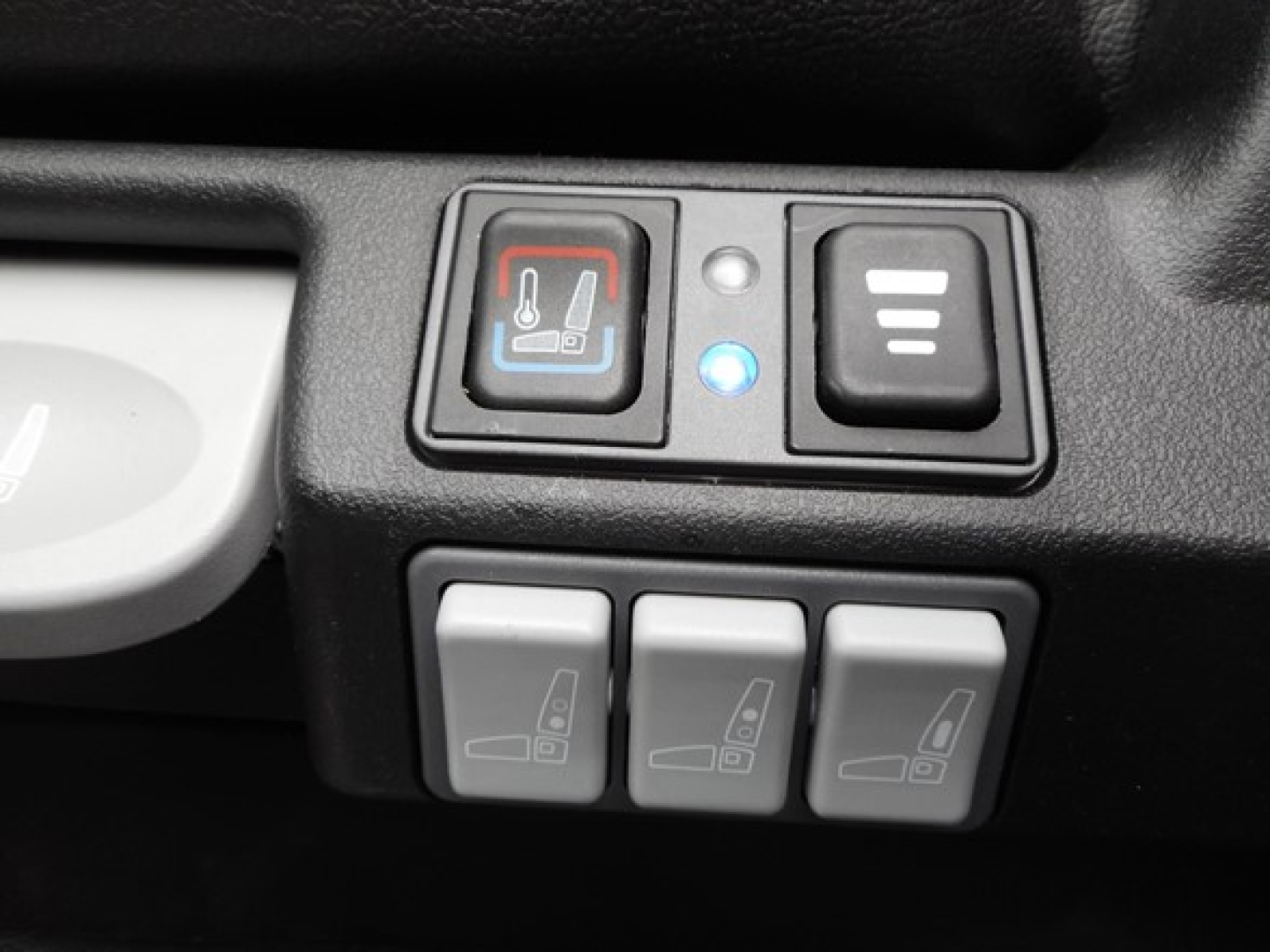 Three gray control switches for seat/mirrors with iconography; a blue backlit indicator and a red/blue seat heating icon on the left; part of a vehicle interior.