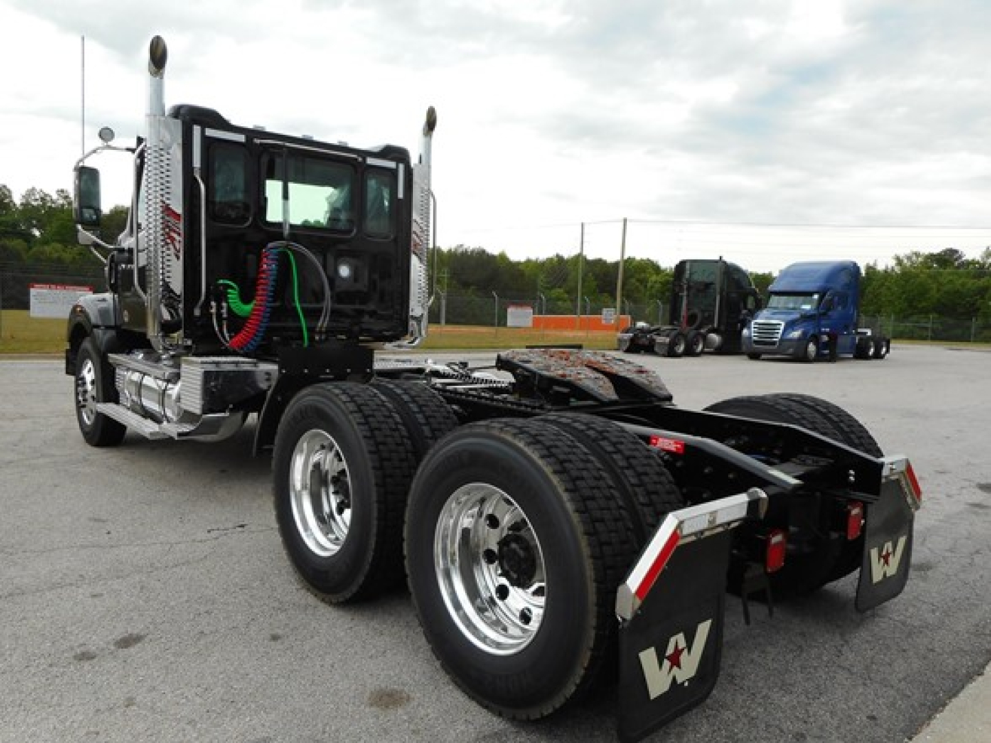 Semi truck chassis in daylight with dual rear axles and chrome exhaust stacks, cab with green hoses, and a blue truck in distance.
