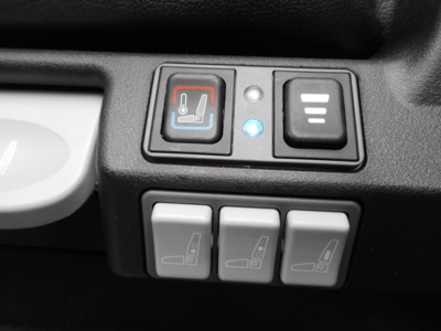 Three gray control switches for seat/mirrors with iconography; a blue backlit indicator and a red/blue seat heating icon on the left; part of a vehicle interior.