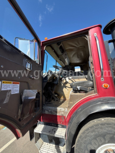 Truck cab interior of a red Mack MR685P, open door showing driver seat and dashboard, stairs visible, blue sky background.