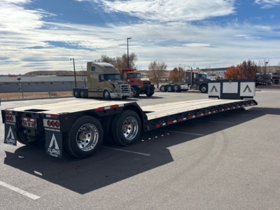 Alpha A80MG lowboy trailer showing long flatbed with dual rear axles and swing-out sides; black and white branding with Alpha trailers logo on mud flaps and rear end.