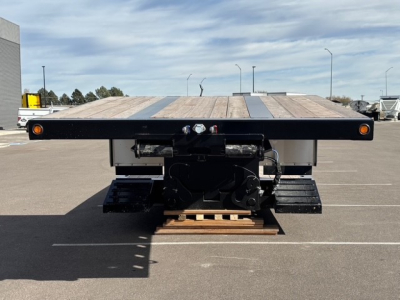 Trailer deck of a lowboy flatbed with swing-out ramps and mechanical detachment mechanism visible from rear; wooden deck, black frame, and undercarriage components.