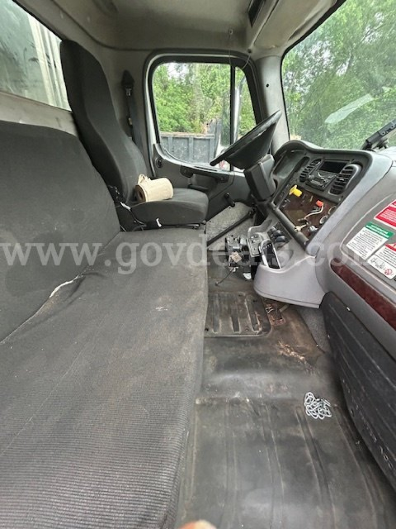 Cab interior of a Freightliner M2 106 with bench seat and dashboard showing steering wheel, instrument cluster, and control panel; worn floor mat and scattered debris visible.