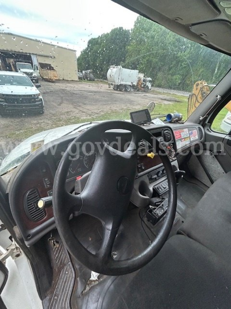 Cab interior of a Freightliner M2 106 showing steering wheel, dashboard with control knobs, and a mounted device; windshield view of yard with trucks and equipment.