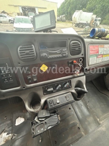 Cab interior of a 2014 Freightliner M2 106 from the driver's side showing dashboard with vents, radio, and switch panel; GPS monitor mounted above; rough cabin with pedals and controls.