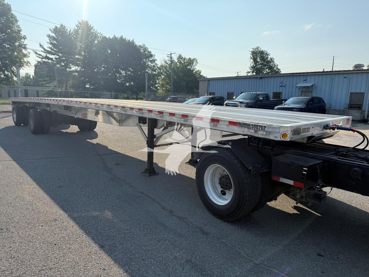 Flatbed trailer showing long aluminum deck with dual rear axles and support legs, side markings, and wheel on the near side.