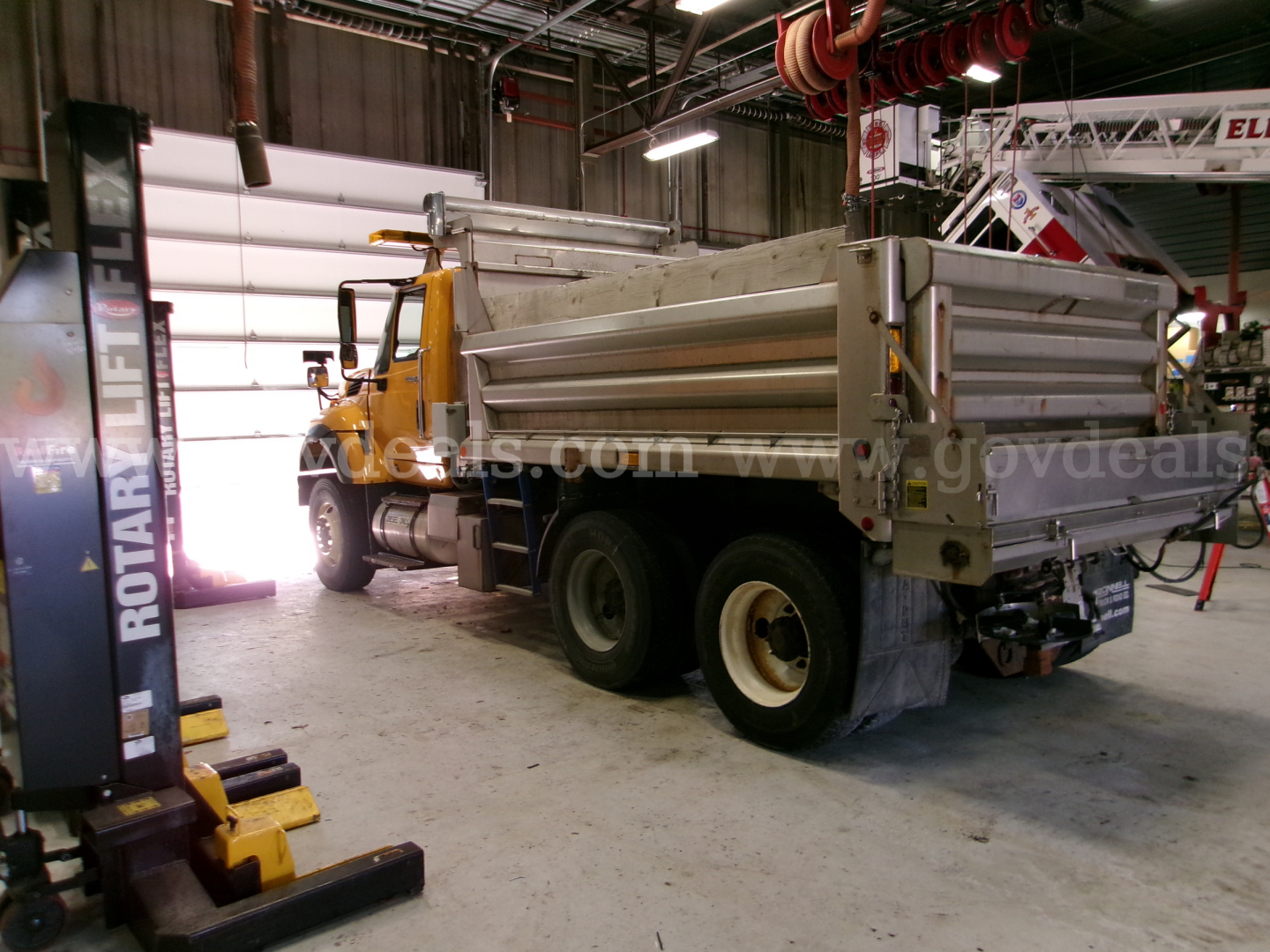 Dump truck chassis and bed of a 2013 International 7400 in a workshop, side view showing cargo bed and dual rear axles with white rims.