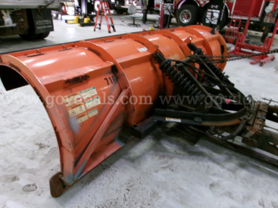 Snowblower attachment or snow plow component in orange with hydraulic hoses and chains, mounted on a truck chassis in a workshop.