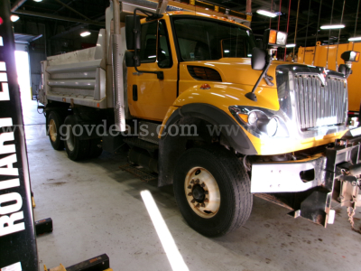 Truck cab with yellow International dump truck in a workshop. Visible text includes 'www.govdeals.com' watermark across the door area and stock/identification labels not fully legible.