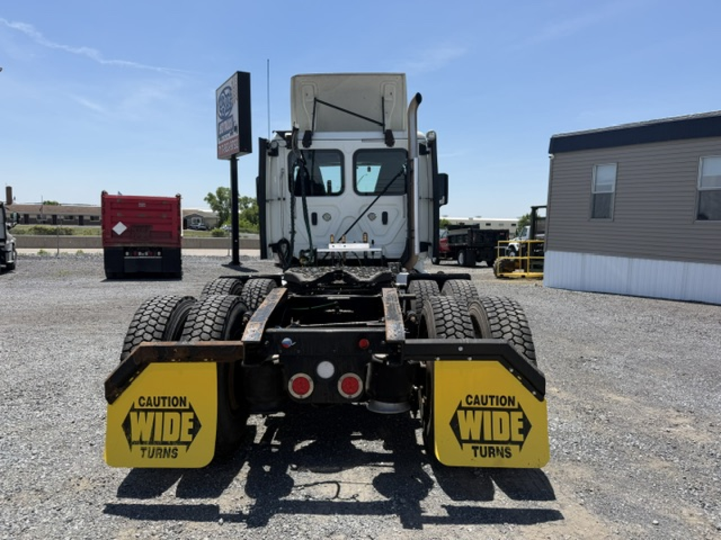 Rear view of a Freightliner Cascadia truck with dual rear axles, yellow caution wide turns mudflaps, highway lot. Visible text: CAUTION WIDE TURNS on mudflaps.