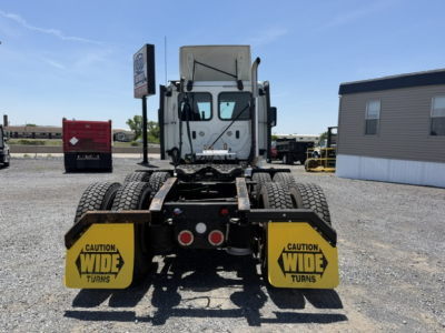 Rear view of a Freightliner Cascadia truck with dual rear axles, yellow caution wide turns mudflaps, highway lot. Visible text: CAUTION WIDE TURNS on mudflaps.