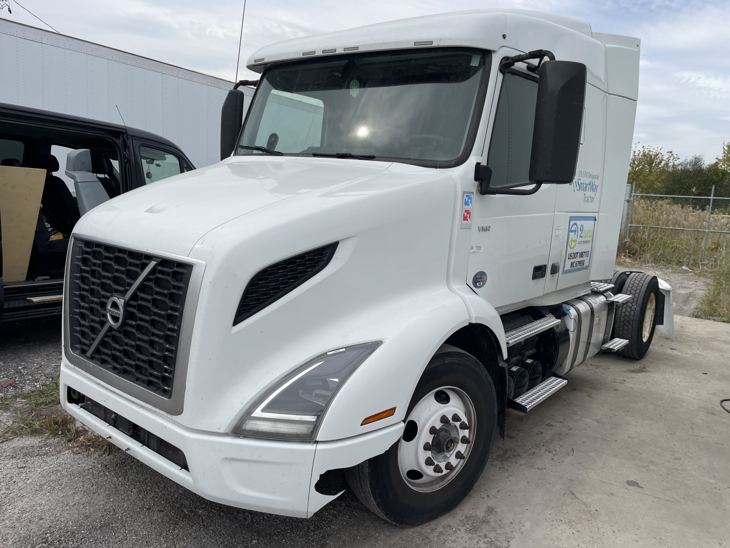 Truck cab showing Volvo VNR day cab with white exterior, large front grille, side mirrors, and company decals on door.