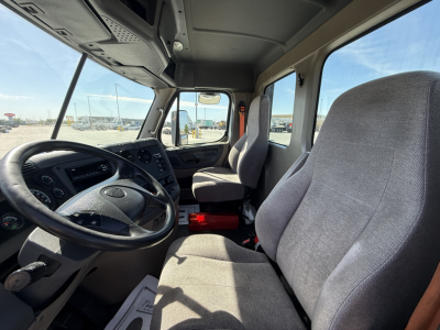 Truck cab interior showing steering wheel and two front seats in a Freightliner CA12564DC with grey upholstery and dashboard controls.