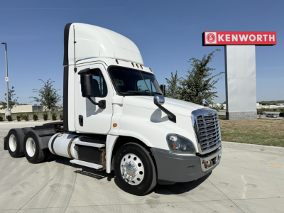 Truck cab and chassis of a white Freightliner CA12564DC Conventional Daycab showing side view with Kenworth sign in background.