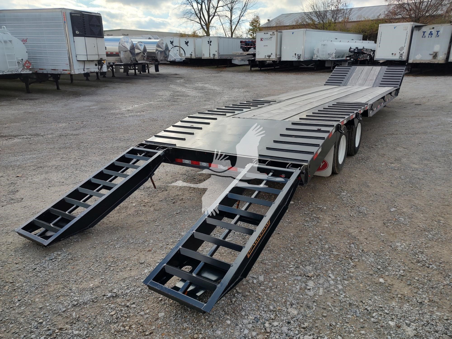 Trailer deck with ramps lowered, multi-axle lowboy trailer in a gravel lot with other trailers in background.