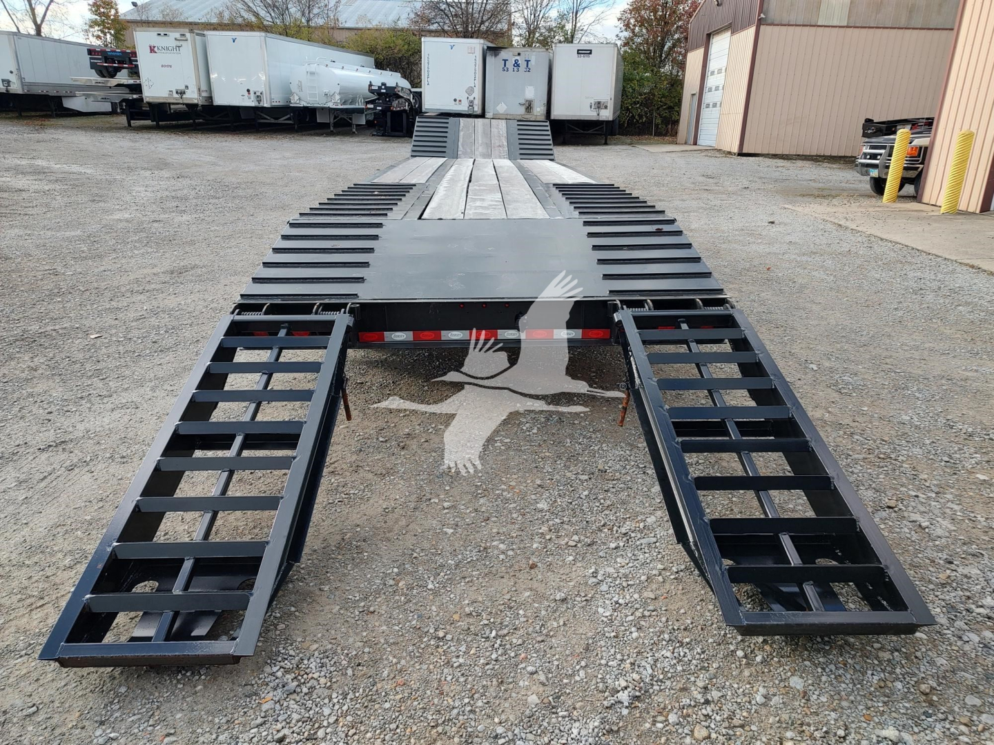 Flatbed trailer showing extendable ramps and deck with black metal frame, trailers in background, gravel ground.