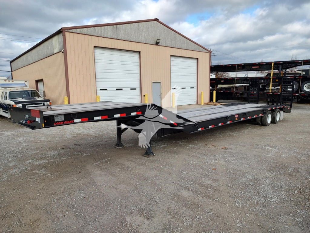 Trailer two-axle lowboy deck showing flatbed surface and front hitch, parked outside a building with overhead doors; stock and VIN not visible in text on image.