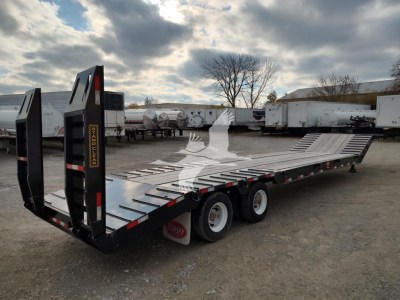 Flatbed trailer with drop deck and rear ramps, black frame, dual rear axles, and multiple crossmembers. Open sky over a gravel lot with other trailers in background.