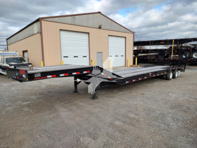 Trailer two-axle lowboy deck showing flatbed surface and front hitch, parked outside a building with overhead doors; stock and VIN not visible in text on image.