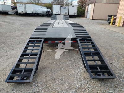 Flatbed trailer showing extendable ramps and deck with black metal frame, trailers in background, gravel ground.