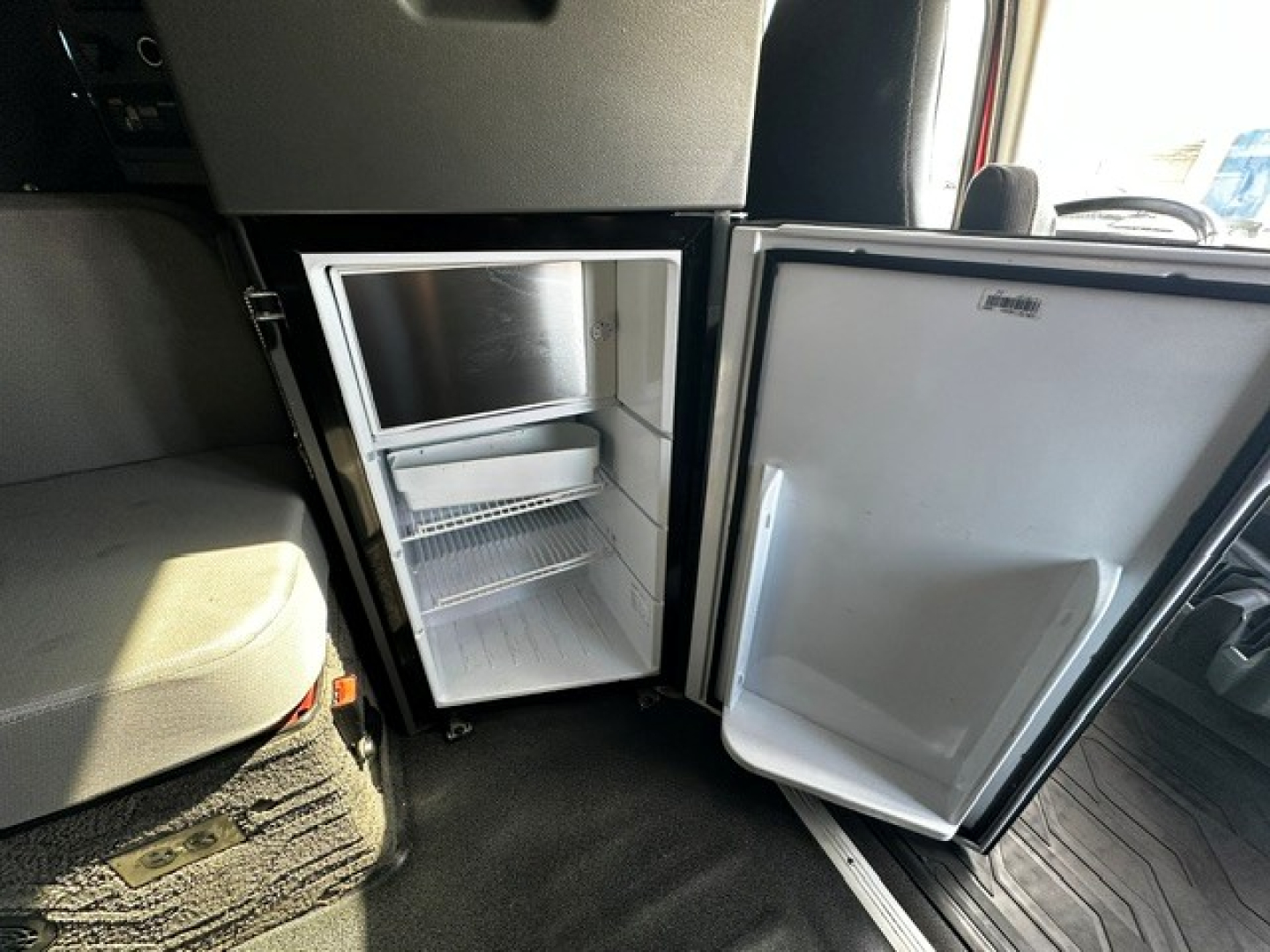 Fridge/freezer unit inside a Volvo VNL64T860 cab with door open showing empty freezer and two shelves, compact refrigerator under dashboard.