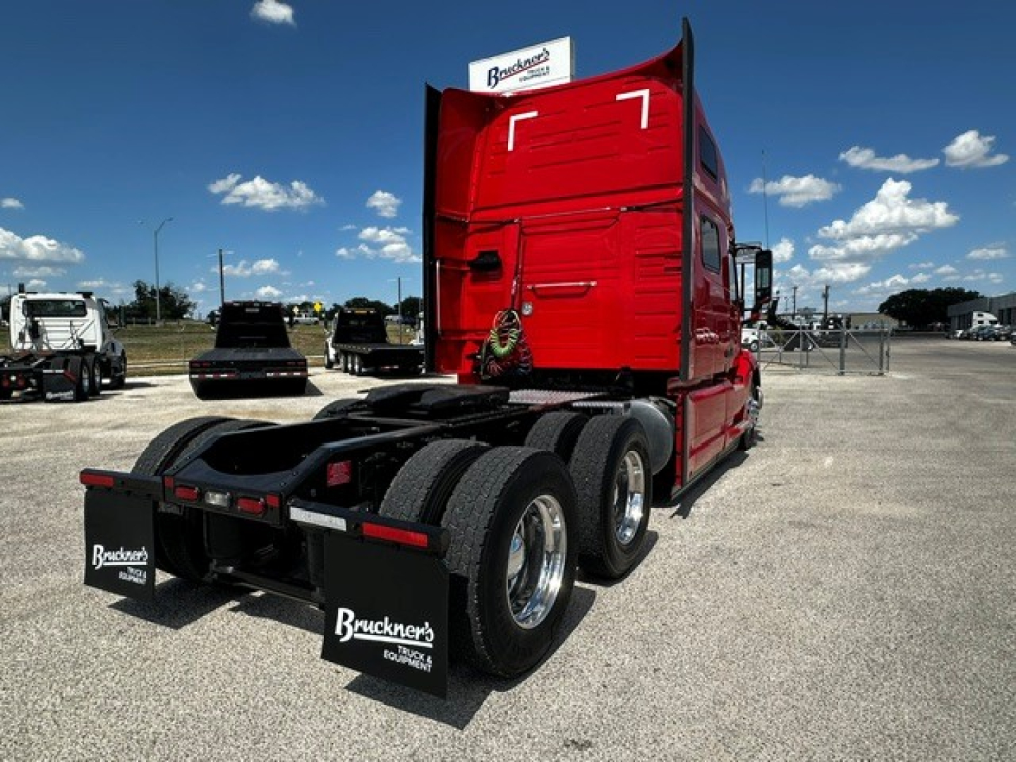 Rear view of red Volvo VNL64T860 sleeper tractor with dual rear axles and mud flaps reading Bruckners Trucking. Clear sky, dealership lot.