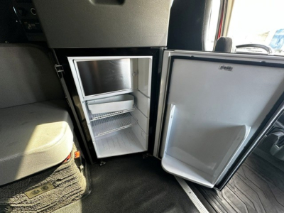 Fridge/freezer unit inside a Volvo VNL64T860 cab with door open showing empty freezer and two shelves, compact refrigerator under dashboard.