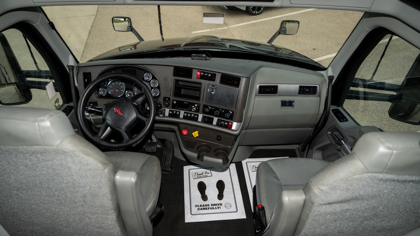 Kenworth T680 cab interior showing steering wheel and instrument cluster with dashboard controls and gray seats.