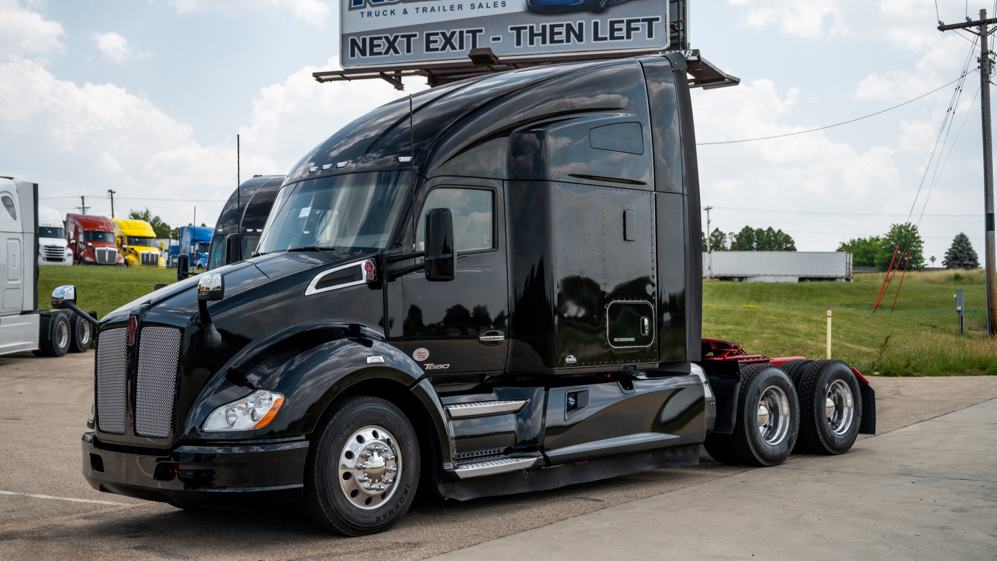 Kenworth T680 truck in a lot showing a sleek black sleeper tractor with three axles and chrome wheels, side view.