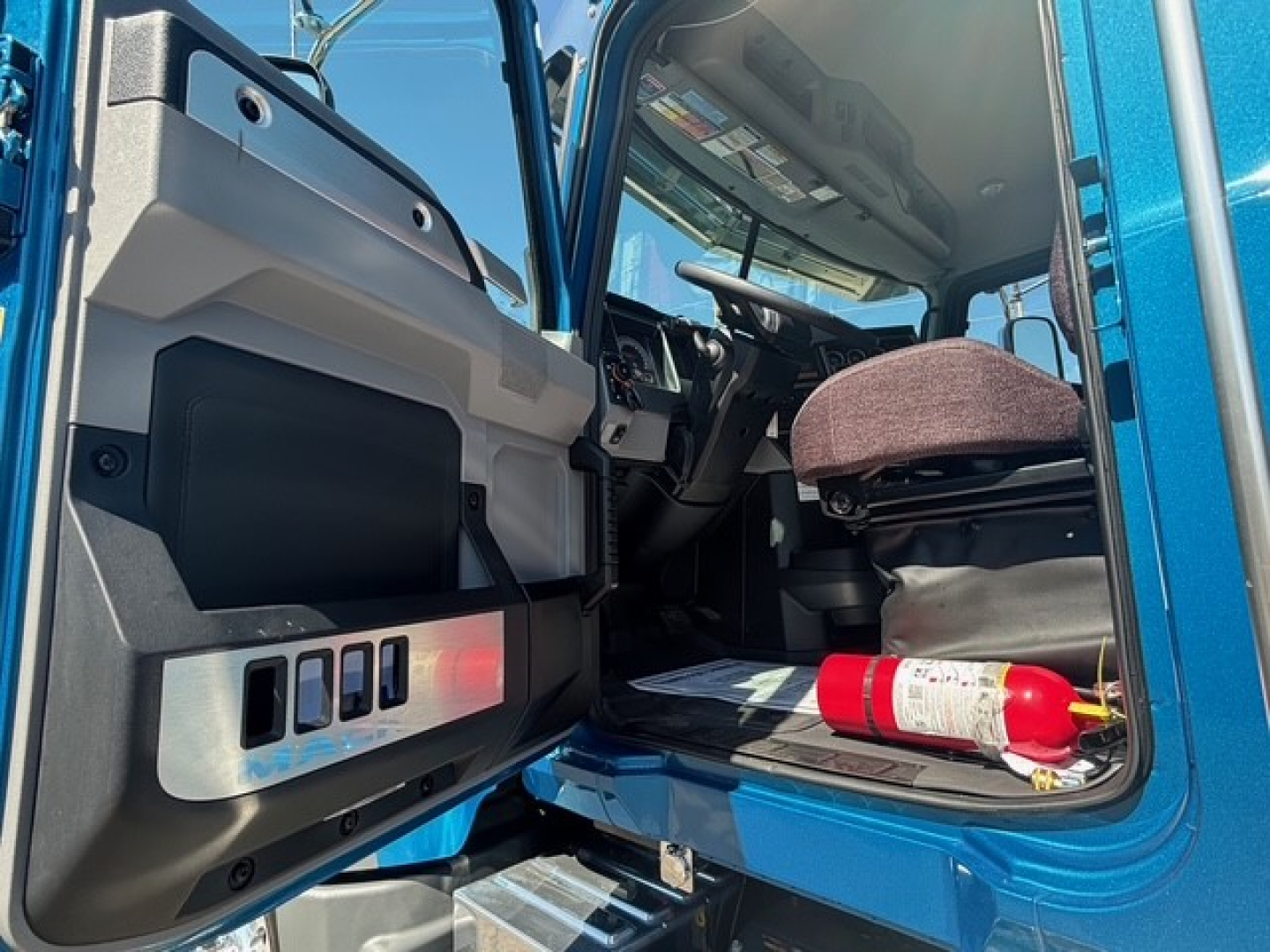Truck cab interior of a 2026 Mack GR64F, focused on the open driver door revealing dashboard controls, seat, and a fire extinguisher in the footwell.
