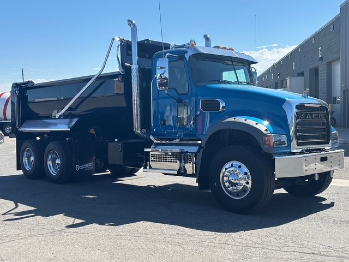 Dump truck with blue cab and black dump body, Mack GR64F, side view showing large chrome bumper, dual rear axles, and hydraulic dump mechanism.