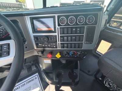 Dashboard view of a Mack GR64F dump truck cab showing multiple gauges, switches, and control panels with steering wheel on left; center stack includes digital display and round gauges.